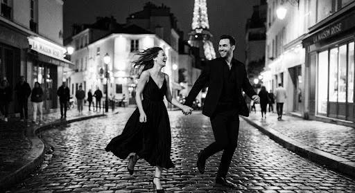A romantic, grainy black and white photograph of a couple running through a Parisian street at night. The city lights are blurred in the background, creating a soft bokeh effect. The couple's joy is palpable as they hold hands, their attire caught in a motion-blur that gives the image a timeless, cinematic quality. The overall mood is one of pure, unrestrained romance and high-fashion elegance.